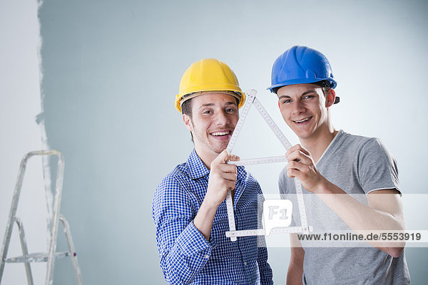 Two happy young men wearing hard hats holding folding rule in shape of a model house