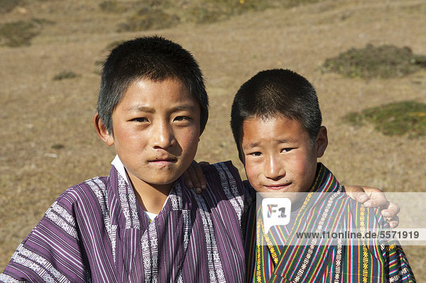 Portrait  zwei Jungs Arm in Arm auf Weide  Tracht  traditionelle Kleidung Gho  bei der Gangtey Goenba  Gangteng Kloster  Phobjika-Tal  Himalaja  Königreich Bhutan  Südasien  Asien