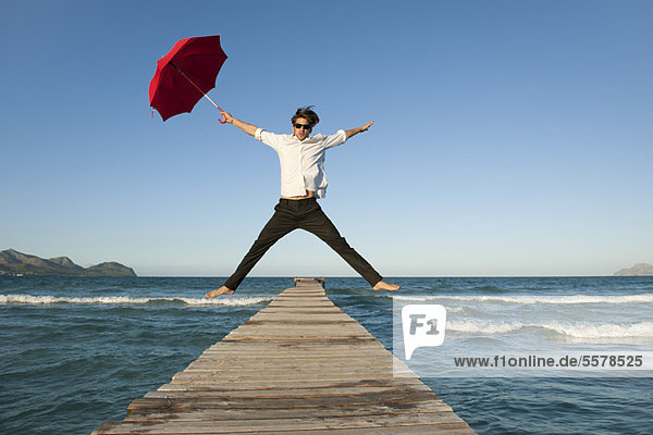 Junger Mann springt auf Pier mit Regenschirm