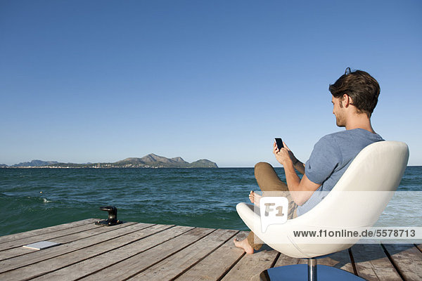 Junger Mann im Sessel am See mit Handy  Seitenansicht