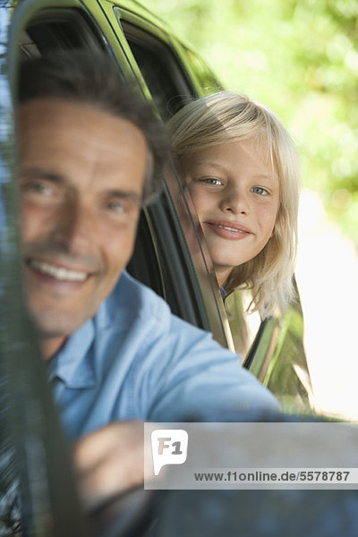 Junge fährt mit Vater im Auto  lehnt sich aus dem Fenster und lächelt in die Kamera.
