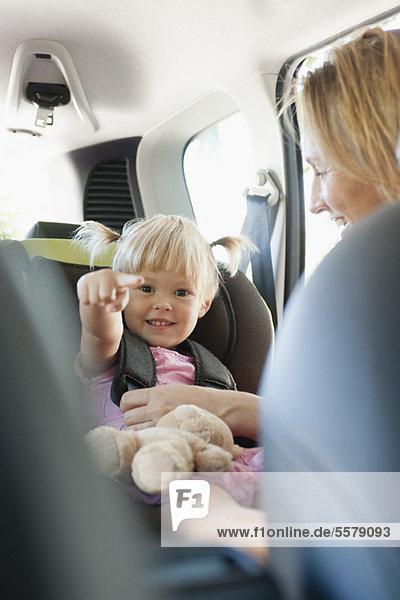 Kleines Mädchen zeigt und lächelt  als ihre Mutter sie im Autositz befestigt.