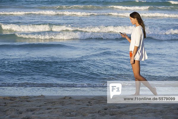 Junge Frau beim Bummeln am Strand mit dem Handy  Seitenansicht