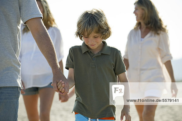 Junge  der mit seiner Familie am Strand spazieren geht und Vaters Hand hält.