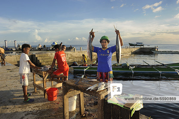 Papua Fischverkäufer hält zwei Fische in die Höhe  die er ausnehmen wird  daneben Kinder  die für Käufer ebenfalls gegen Geld Fische ausnehmen  auf dem Fischmarkt in Kota Biak  Insel Biak  West-Papua  Indonesien  Südostasien  Asien