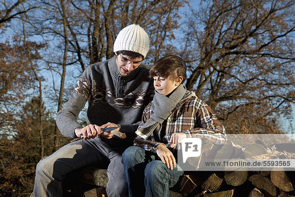 Germany  Bavaria  Father and son sitting on wood and cutting stick