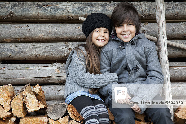 Girl and boy sitting on stack of firewood  smiling  portrait