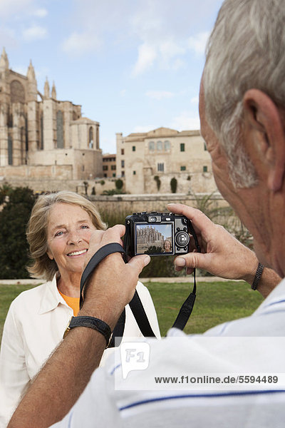 Spanien  Mallorca  Palma  Seniorenpaar lächelnd beim Fotografieren mit der Kathedrale Santa Maria