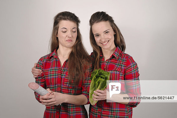 Zwillingsschwestern  die eine mit einer Salami in der Hand  die andere mit einem Salatkopf