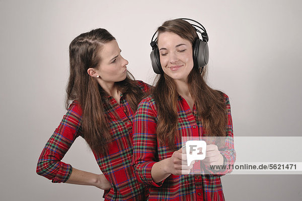 Twin sisters  one holding an iPod and listening to headphones  the other looking enviously