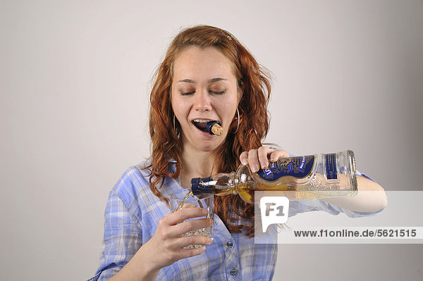 Junge Frau mit roten Haaren öffnet eine Flasche Whiskey mit den Zähnen und gießt sich ein Glas ein