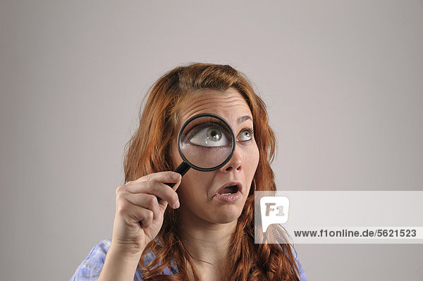 Young woman with red hair looking with one eye through a magnifying glass