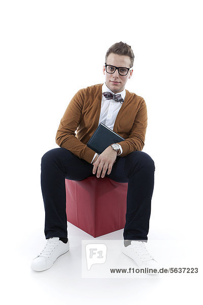 Junger Mann mit Brille und Fliege hält Buch  Agenda und sitzt auf rotem Sitzwürfel