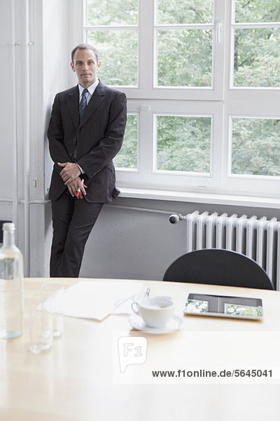 Ein Geschäftsmann  der sich gegen eine Fensterbank in einem Konferenzraum lehnt.