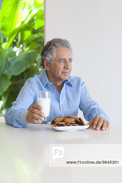 Ein älterer Mann sitzt an einem Tisch mit Schokokeksen und einem Glas Milch.