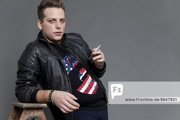 Junger Mann in Lederjacke und Jeanshose raucht Zigarette und stützt sich lässig auf alte Holzleiter