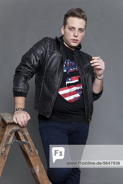 Junger Mann in Lederjacke und Jeanshose raucht Zigarette und stützt sich lässig auf alte Holzleiter