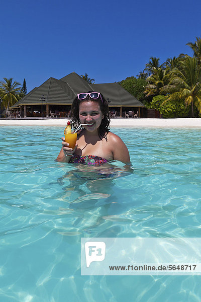 Mädchen  ca. 14 Jahre  mit Sonnenbrille im Haar  trinkt einen Cocktail in einer türkisfarbenen Lagune im Meer  hinten Malediveninsel Paradise Island  Lakanfinolhu  Malediven  Indischer Ozean  Asien