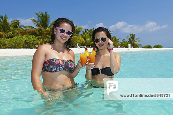 Zwei Mädchen  ca. 14 und 18 Jahre  mit Sonnenbrillen  trinken Cocktails in einer türkisfarbenen Lagune im Meer  hinten Malediveninsel  Malediven  Indischer Ozean  Asien