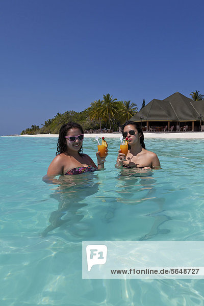 Zwei Mädchen  ca. 14 und 18 Jahre  mit Sonnenbrillen  trinken Cocktails in einer türkisfarbenen Lagune im Meer  hinten Malediveninsel Paradise Island  Lakanfinolhu  Malediven  Indischer Ozean  Asien