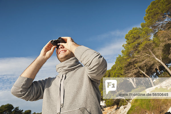 Spain  Mallorca  Young man looking through binocular  smiling