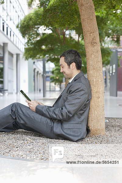 Germany  Leipzig  Businessman sitting at tree and using digital tablet