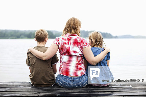 Rückansicht der Mutter mit Arm um ihre Kinder auf dem Pier sitzend