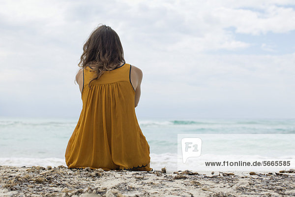 Junge Frau am Strand sitzend mit Blick auf den Strand  Rückansicht