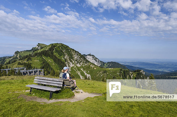 Frau sitzt auf einer Holzbank und genießt den Blick vom Brauneckgipfel  Bayern  Deutschland  Europa