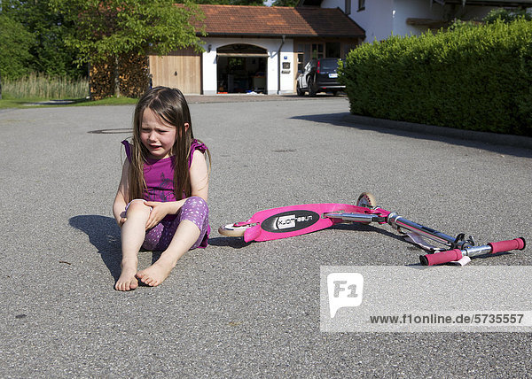 Dreijähriges Mädchen ist mit Roller hingefallen  sitzt weinend auf dem Boden und hält sich das verletzte Knie
