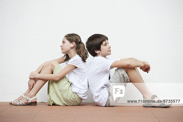Junge und Mädchen sitzen Rücken an Rücken und hören zusammen Musik.