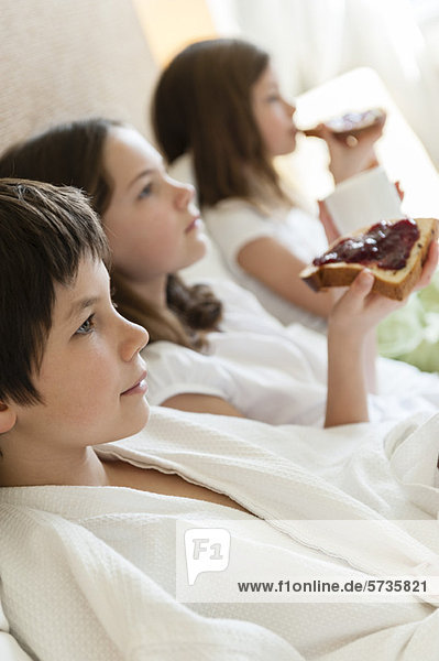Geschwister entspannt im Bett  Mädchen essen Toasts im Hintergrund
