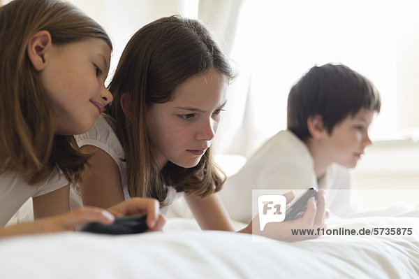 Sisters lying on bed playing handheld video game  boy in background