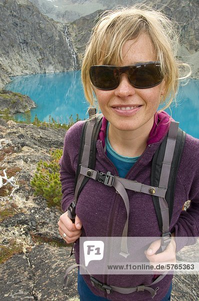 Eine junge  attraktive Frau mit Sonnenbrille lächelt vor einem türkis blaue See und Wasserfall in der Coast Mountains  British Columbia  Kanada