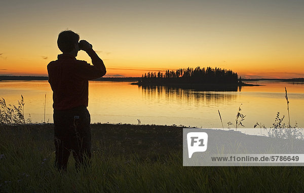 Middle age male hiker looking out over Astotin Lake with binoculars at dusk  Elk Island Park  Alberta  Canada.