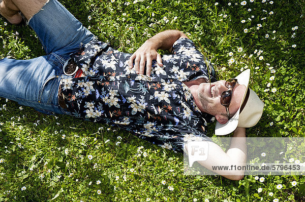 Lächelnder Mann mit Strohhut  Sonnenbrille und Hawaiihemd liegt entspannt in einer Blumenwiese und hat einen Grashalm im Mund