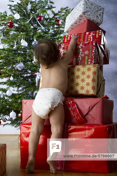 Baby Mädchen steht auf Zehenspitzen  um einen hohen Stapel Weihnachtsgeschenke zu erreichen.