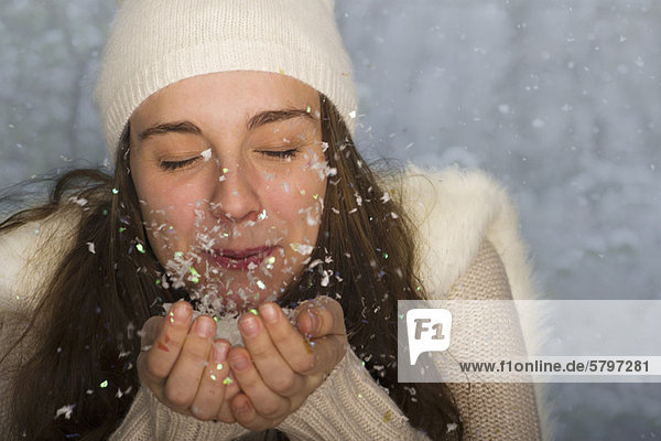 Junge Frau in Winterkleidung  bläst eine Handvoll Konfetti  Mund