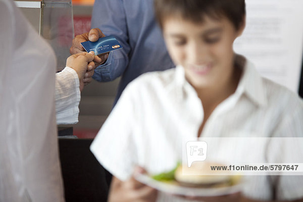 Frau  die für den Kauf mit Kreditkarte bezahlt  Junge  der einen Teller mit Essen im Vordergrund trägt.
