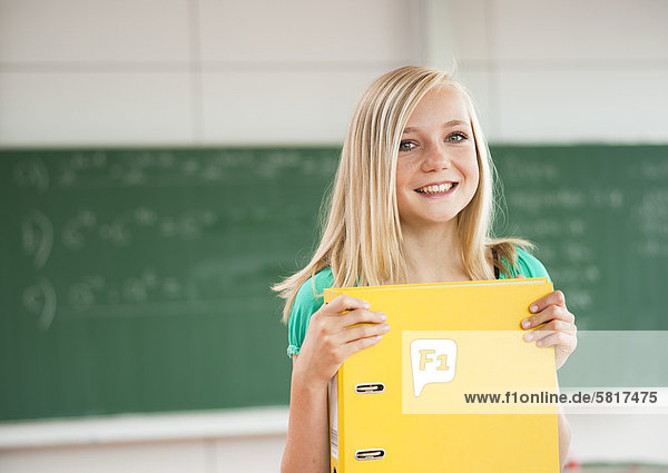 Lächelnde Teenagerin im Klassenzimmer hält einen Aktenordner  Portrait