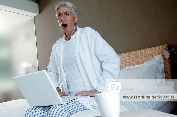 Senior man sitting on the bed with a laptop on his lap