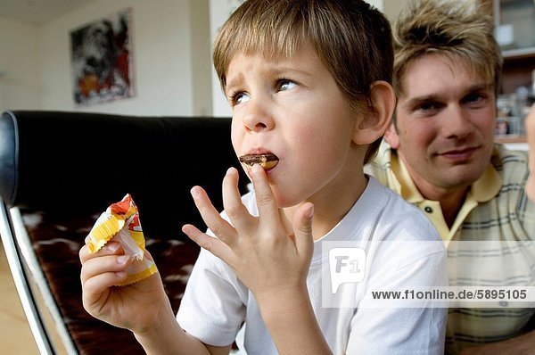 Close_up of a boy eating a chocolate bar with his father sitting behind him