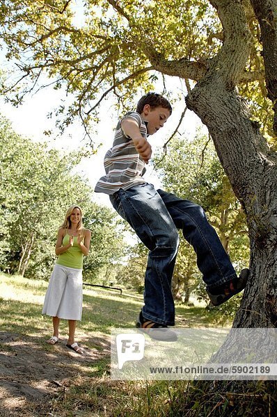 Side profile of a boy climbing a tree with his mother clapping in a park