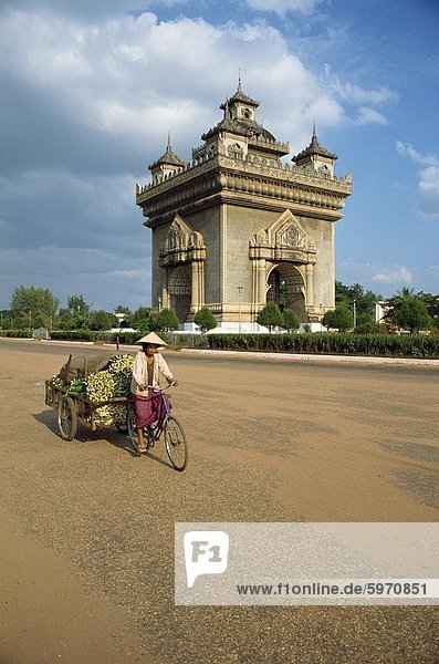 Frau auf einem Fahrrad mit Anhänger fährt vorbei an das Anousavari-Denkmal in Vientiane  Laos  Indochina  Südostasien  Asien