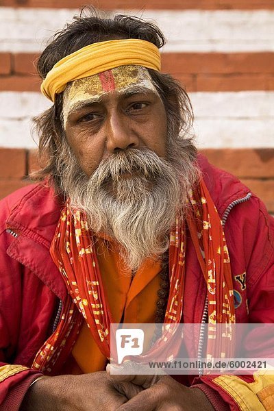 Ein bärtiger Sadhu trägt ein Halstuch und sitzt durch rote und weiße Ghats in Varanasi Uttar Pradesh Indien Asien