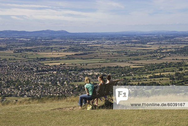 Drei Mädchen auf Bank betrachten die Ansicht über Bishops Cleeve Dorf  Cleeve Hill  The Cotswolds  Gloucestershire  England  Vereinigtes Königreich  Europa