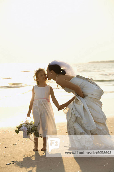 Caucasian mid-adult bride kneeling to give flower girl a kiss on the cheek while holding hands barefoot on beach.