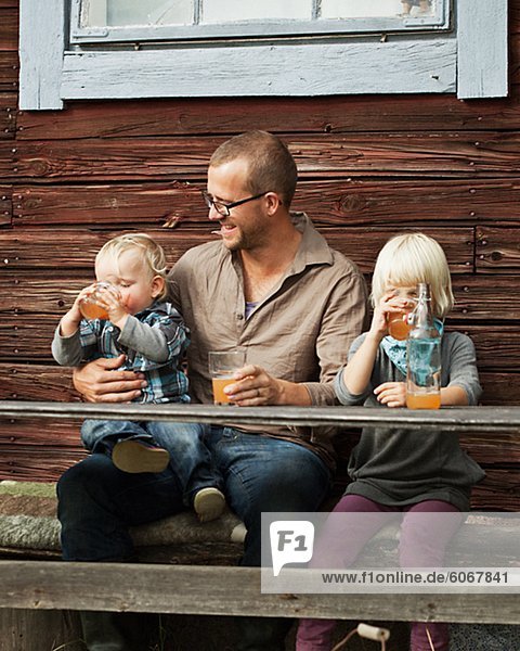 Father with two kids drinking juice in front of wooden house