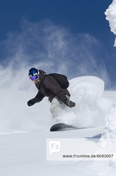 A young man backcountry snowboards in deep powder against a blue sky.