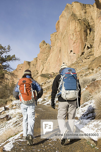 Two rock climbers with backpacks walking down a trail with a cliff in the background.